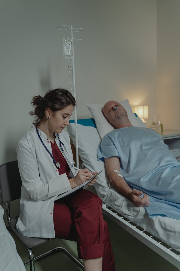 A doctor sits beside a patient in a hospital room, taking notes.