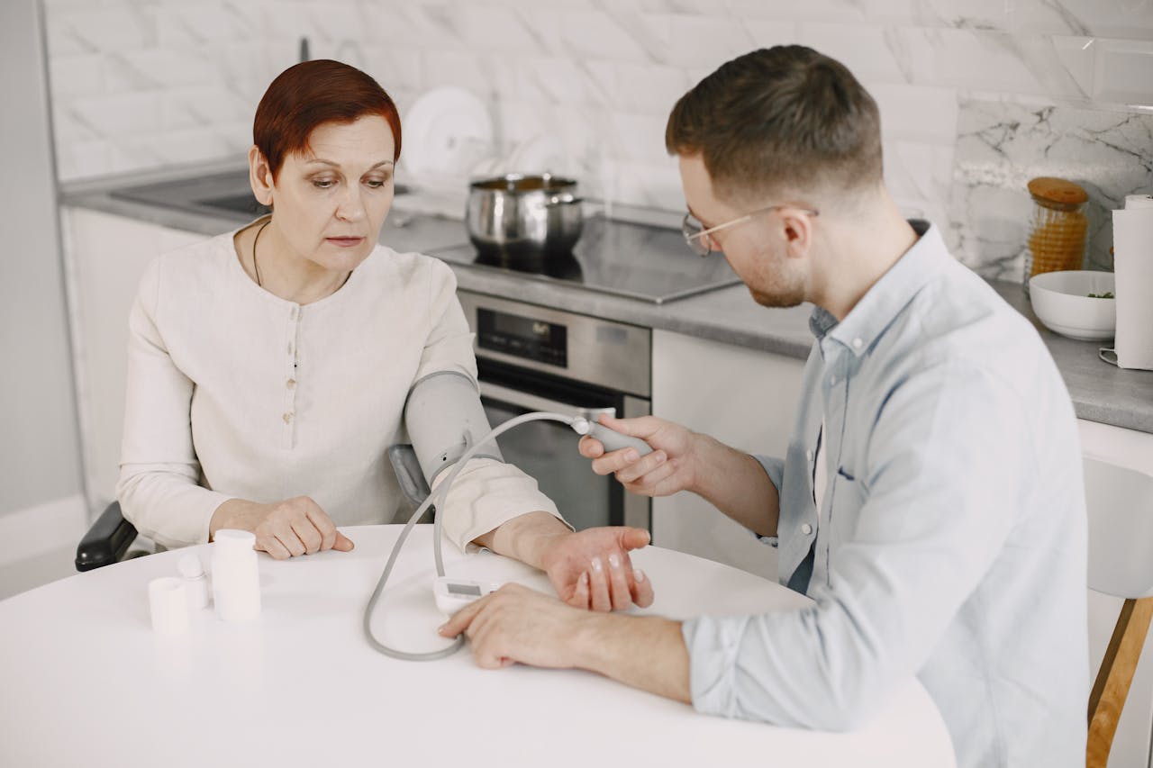 Adult man monitoring elderly woman's blood pressure at kitchen table using sphygmomanometer.